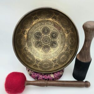 Bol tibétain 7 métaux gravé vu de face, intérieur orné du mantra Om Mani Padme Hum en caractères tibétains, entouré de motifs circulaires finement gravés, posé sur un coussin, avec mailloche feutre et bâton bois-cuir visibles au premier plan