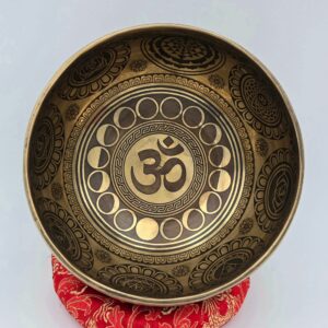 Bol tibétain 7 métaux gravé Om vu de l’intérieur, centre orné du symbole Om entouré des phases lunaires et de motifs géométriques traditionnels, posé sur coussin rouge
