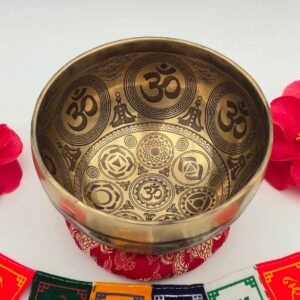 Bol tibétain 7 métaux entièrement gravé – note Sol – 130 mm – vue intérieure avec symboles Om, posé sur coussin rouge avec drapeaux de prière tibétains en arrière-plan