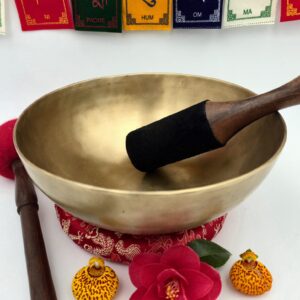 bol chantant tibétain thérapeutique 7 métaux vue intérieure et extérieure avec mailloche bois cuir, bâton et coussin rouge, fleurs décoratives et drapeaux de prière mantra om mani padme hum en arrière-plan