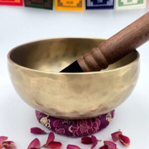 bol tibétain mat 7 métaux 550 g vue extérieure avec bâton bois cuir posé sur coussin violet et pétales de fleurs, drapeaux de prière en arrière-plan